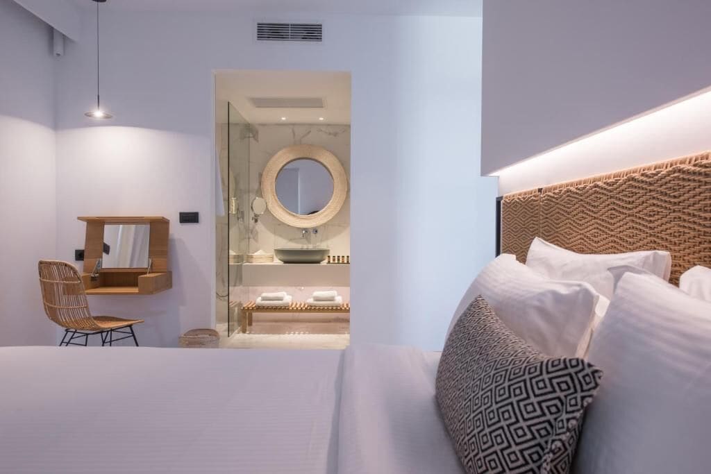 A modern bedroom with a large bed, a wooden headboard, a wicker chair, a bedside table, and a bathroom visible through a glass door.