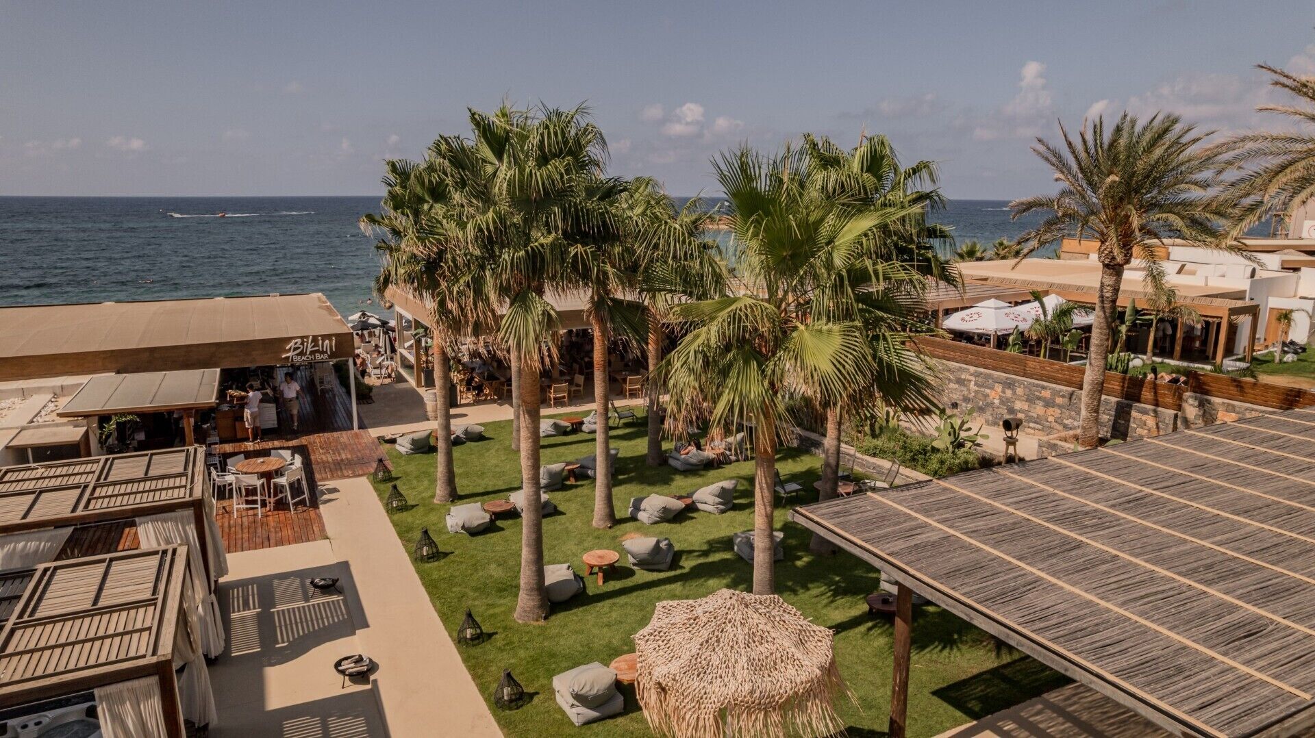 Un resort frente al mar con bar, mesas al aire libre, palmeras y vistas al océano.