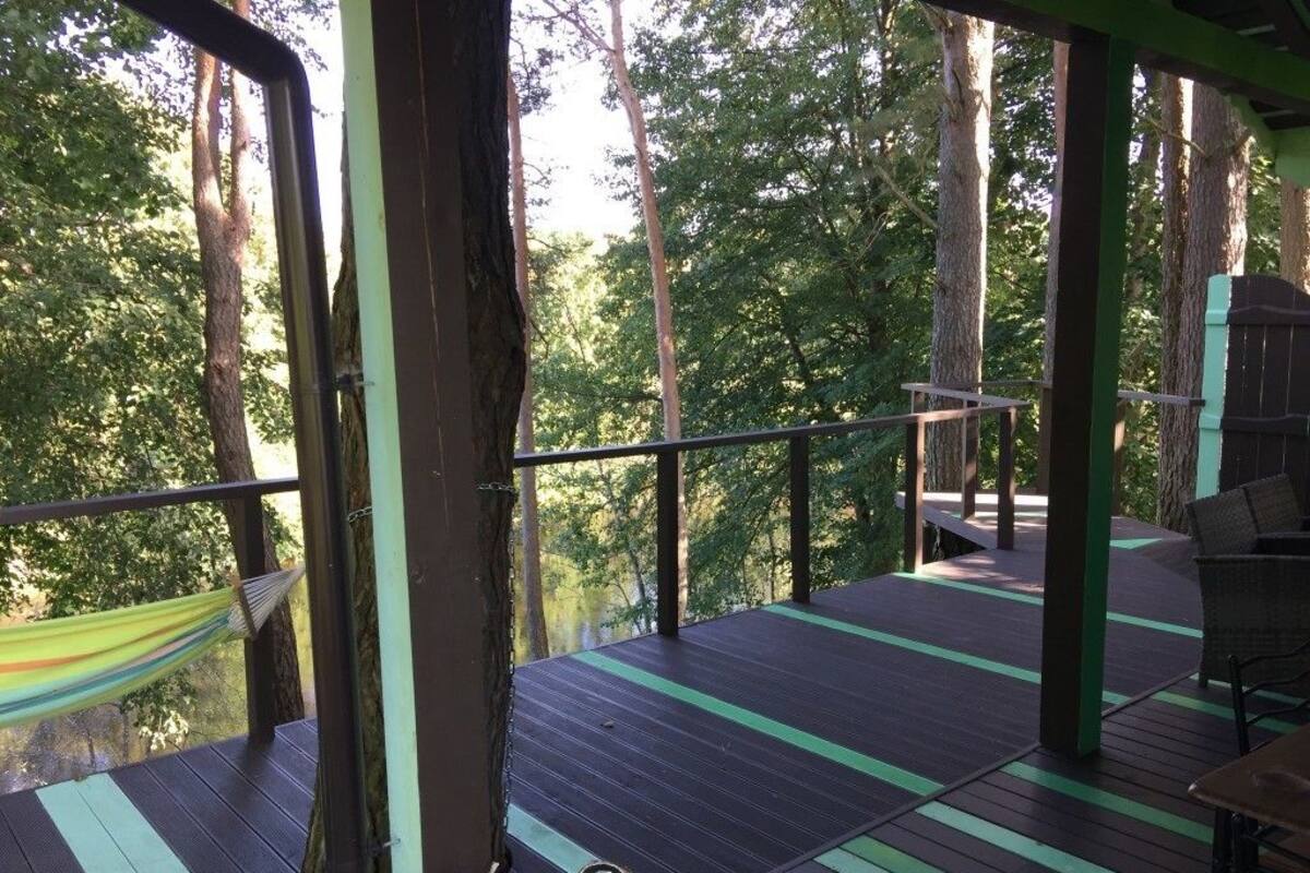 Terraza al aire libre con una hamaca, muebles de madera y barandilla con vistas a un área boscosa.