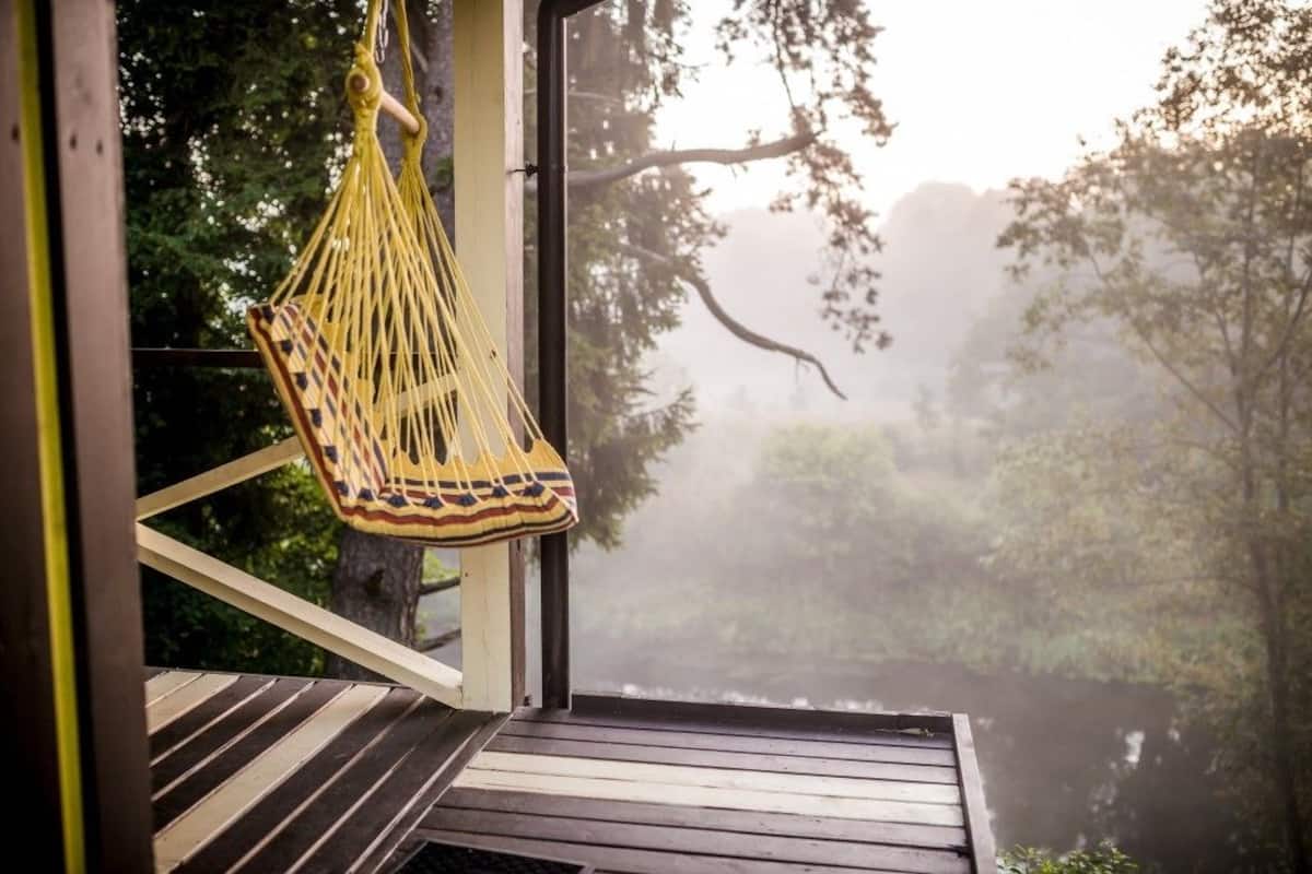 Terraza de madera con una hamaca amarilla colgada y vista a un área boscosa a través de una ventana.