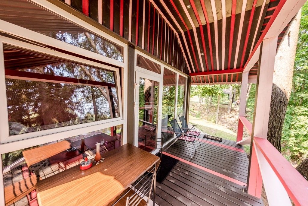 Een overdekt terras met een houten tafel en stoelen, grote ramen en uitzicht op de omliggende begroeiing.