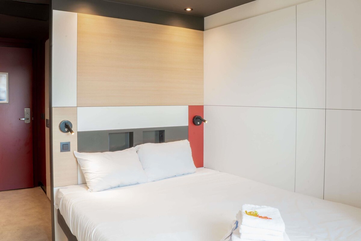 A modern hotel room with a bed, a bedside table, and a wall-mounted control panel.