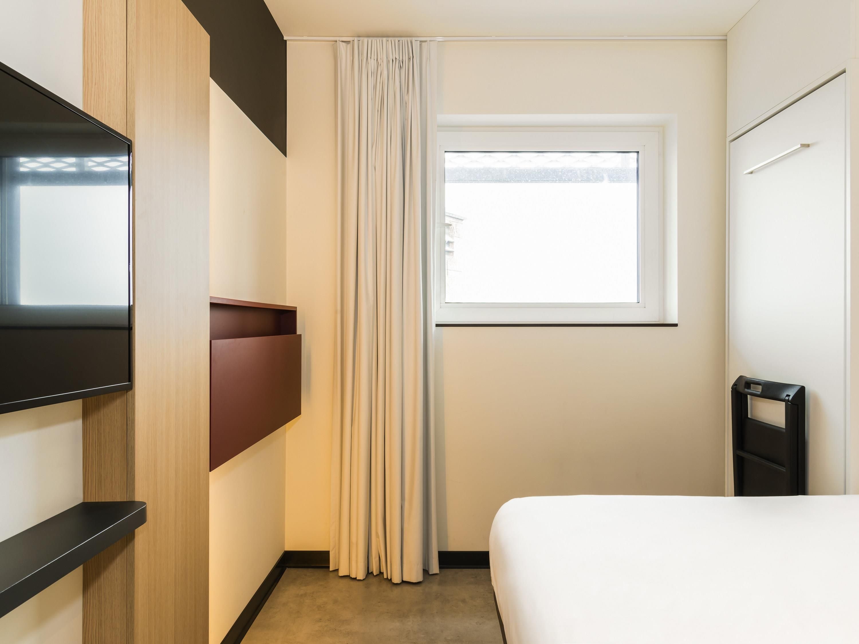 A hotel room with a bed, a TV mounted on the wall, a window with curtains, and a door with a handle.