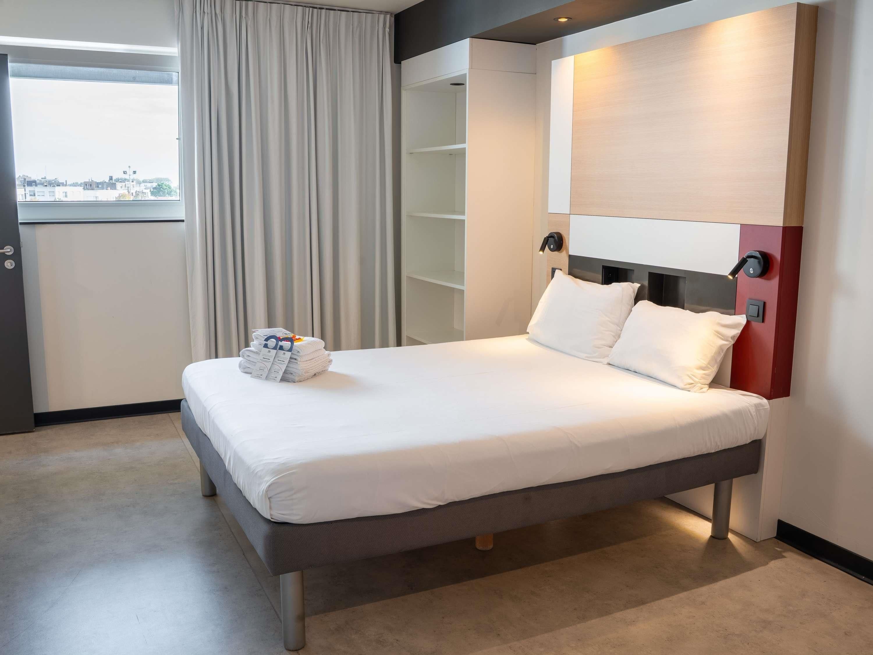 Een hotelkamer met een bed, nachtkastje, gordijnen en uitzicht op de stad.