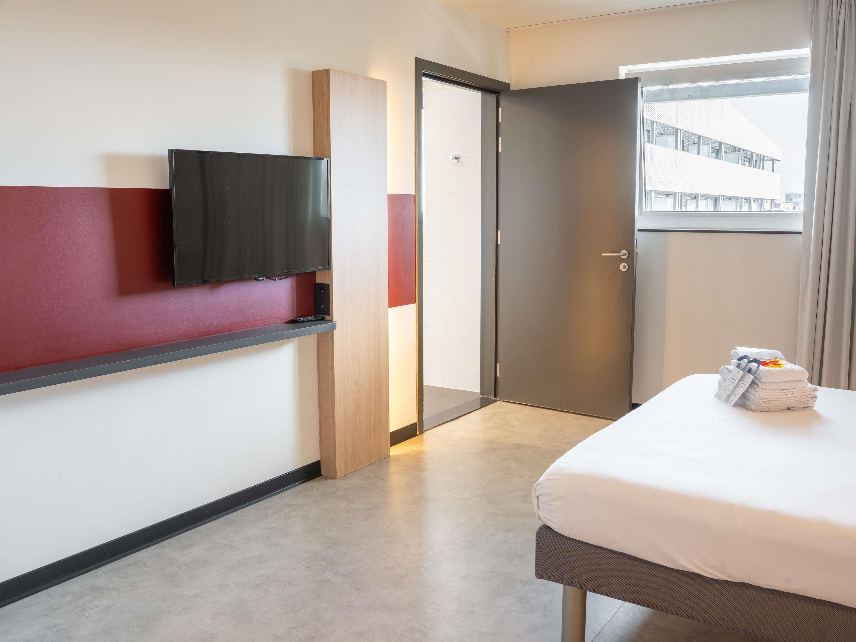 A hotel room with a bed, a flat-screen TV mounted on the wall, a door, and a window with a view of a building.