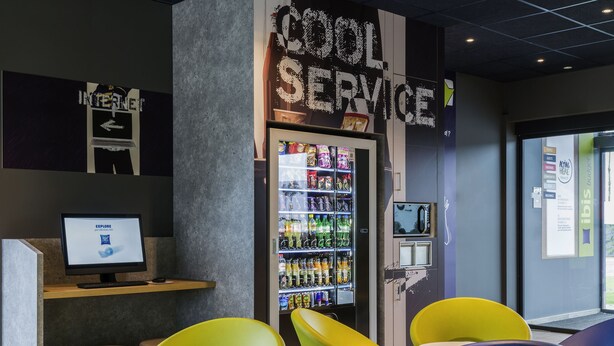 A modern lounge area with colorful chairs, a vending machine, and a computer monitor displaying a logo. - ibis budget Blankenberge (Blankenberge)