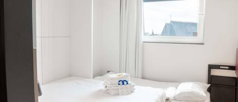 A hotel room with a bed, a window with a view of buildings, a small table, and a chair.