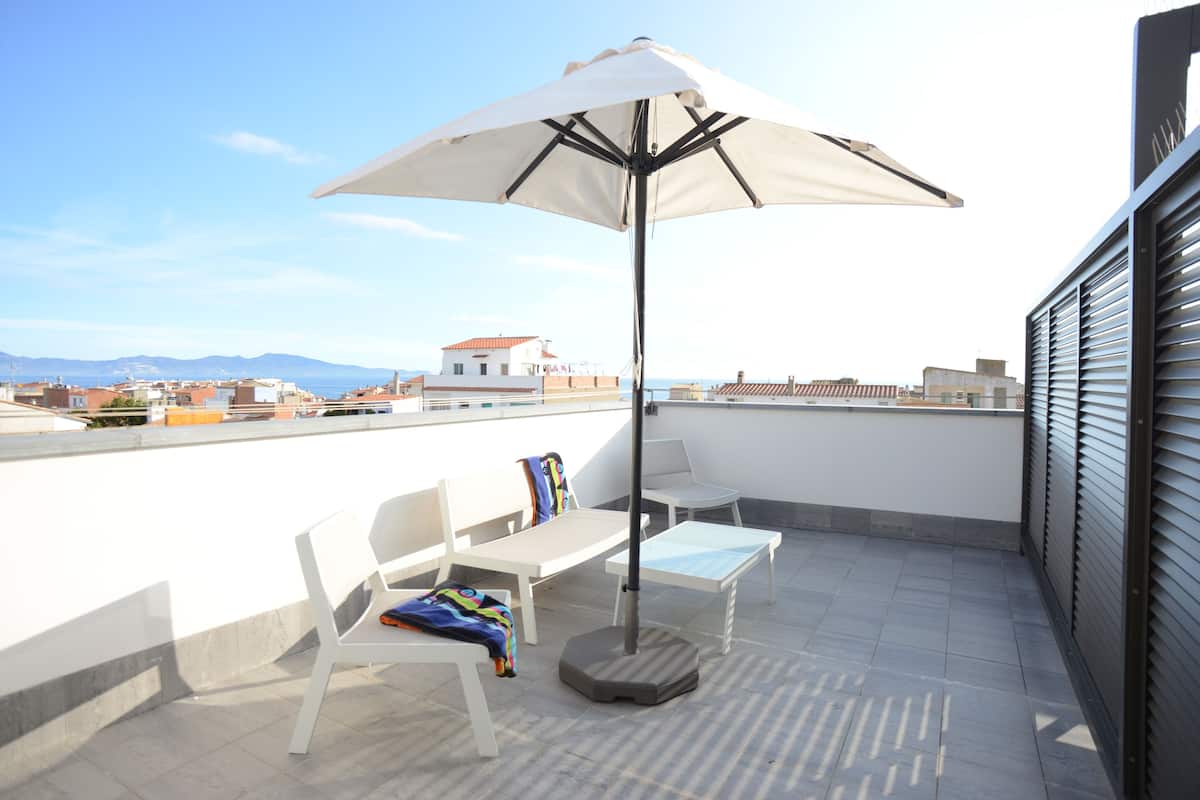 Terraço no topo do prédio com um guarda-sol branco, duas cadeiras brancas e uma mesa pequena.