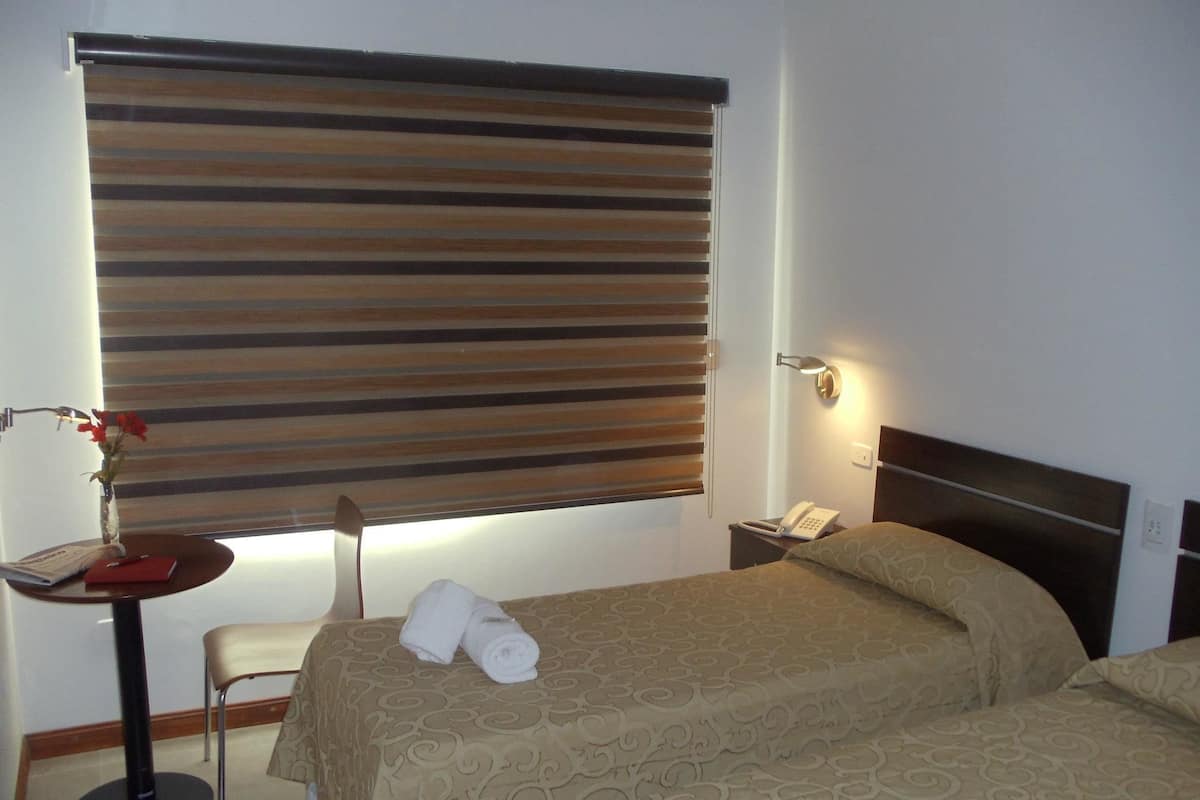 A hotel room with a large window covered by blinds, a bedside table with a lamp, a telephone, and a bed with two pillows and a blanket.