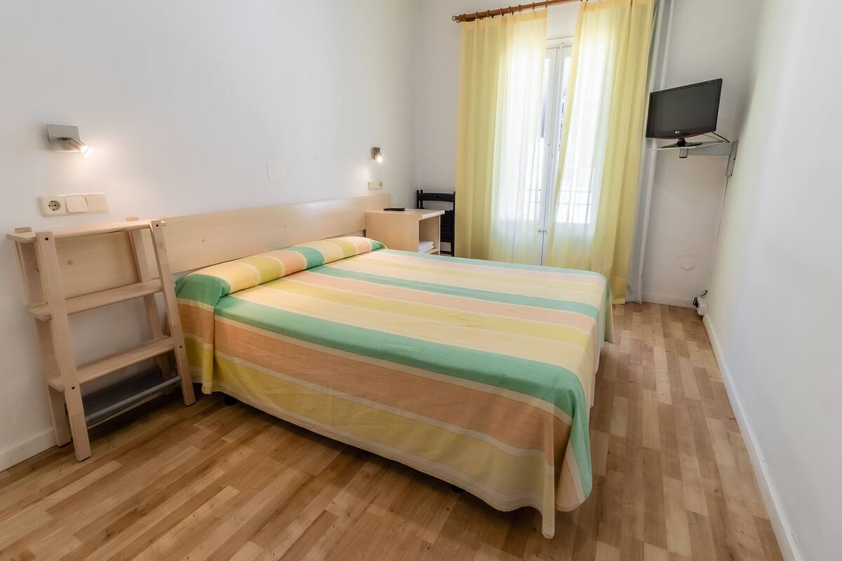 Ein ordentlich eingerichtetes Schlafzimmer mit einem Bett, Nachttischen, einem Fernseher und einem Fenster mit Vorhängen.