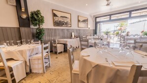Un restaurant avec des tables rondes dressées pour le dîner, des nappes blanches et une vue sur la mer grâce à de grandes fenêtres.