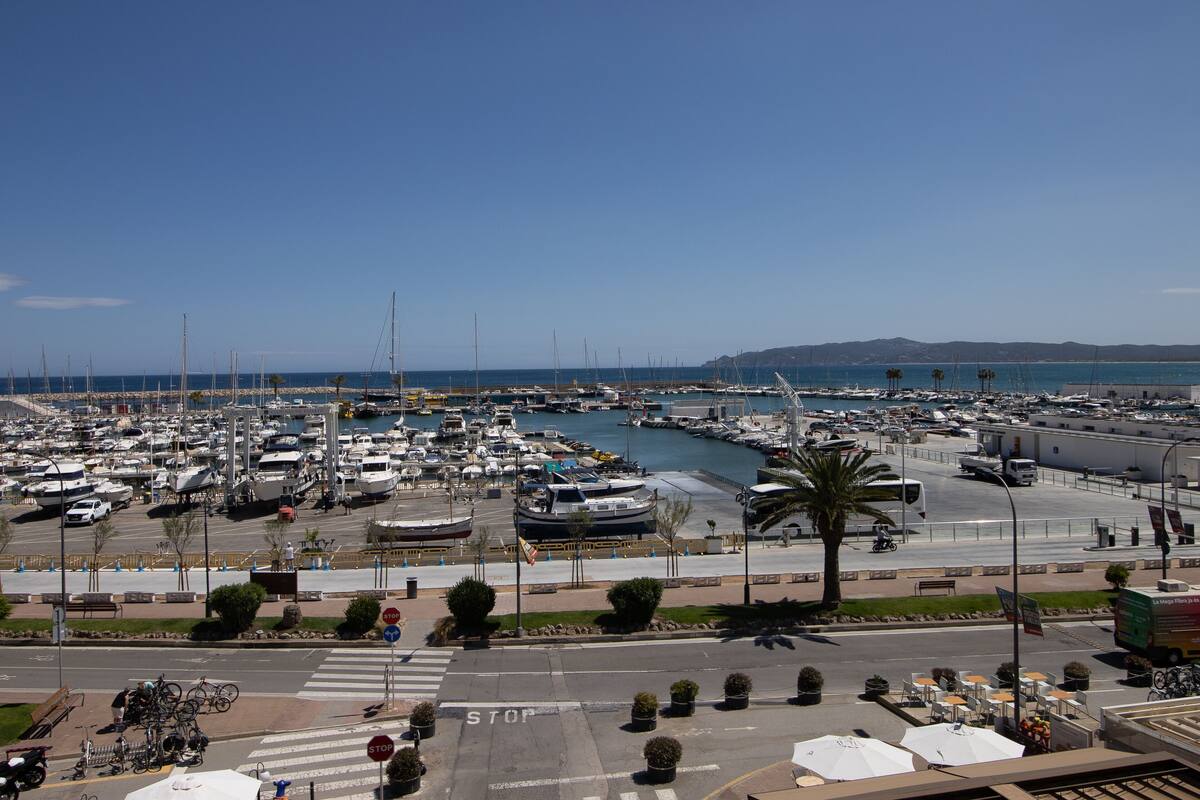 Ein Yachthafen mit vielen vertäuten Booten, einer Fußgängerzone und freiem Blick auf das Meer.