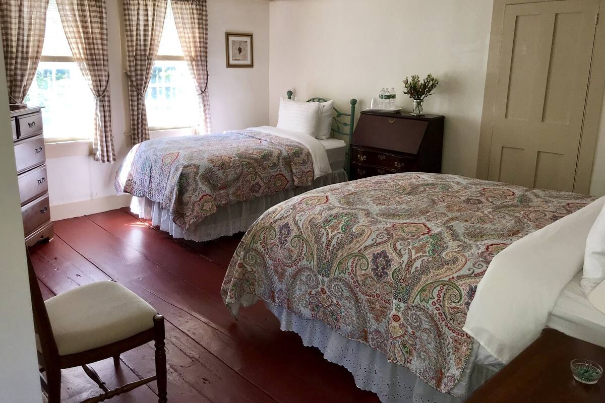 Une chambre avec deux lits simples, une commode en bois, une chaise et une fenêtre avec des rideaux.