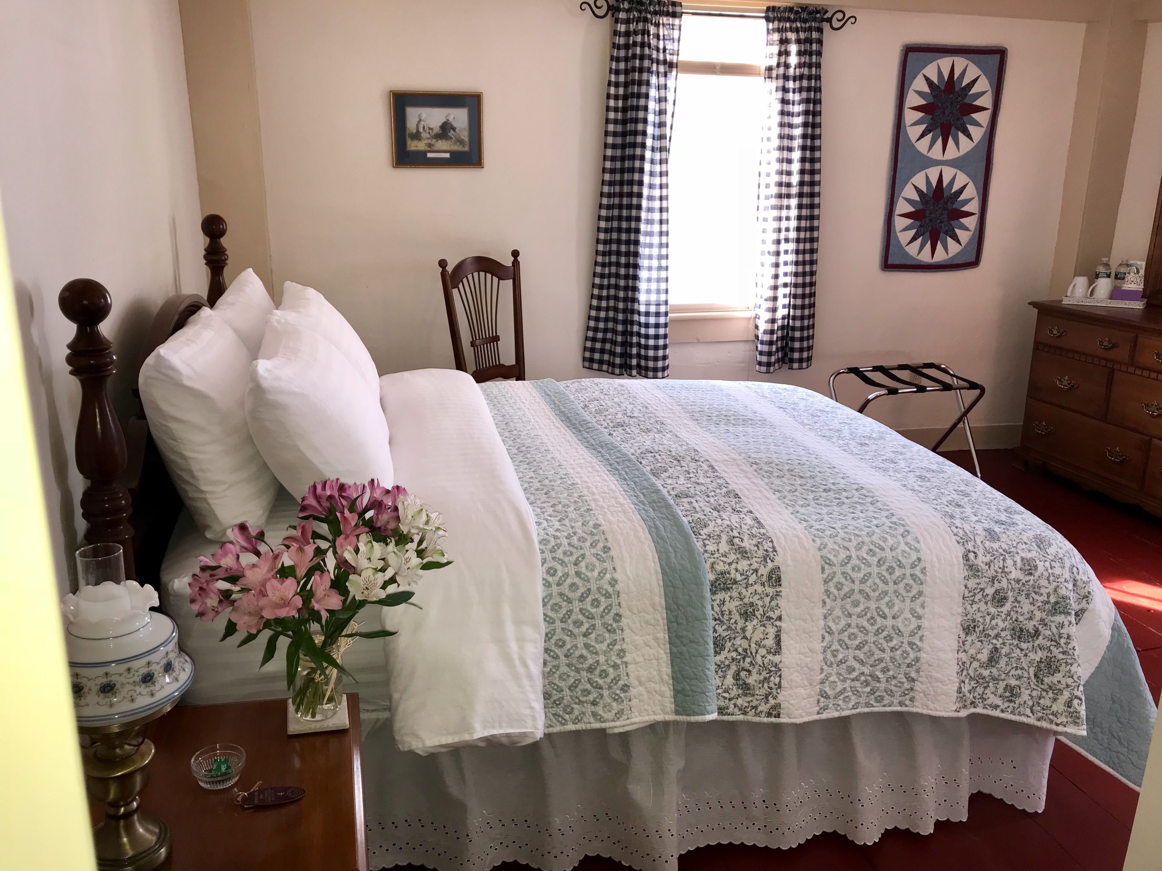 Una cama bien tendida con un arreglo floral, una silla de madera, un aparador y una ventana con cortinas a cuadros.