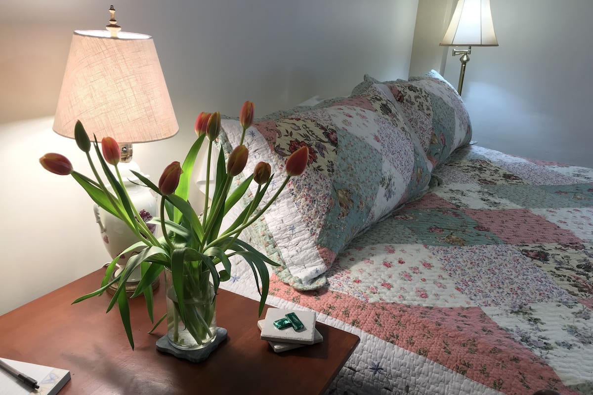 Un lit recouvert d’une courtepointe à motifs floraux, une lampe de chevet, un vase de tulipes et un bloc-notes posés sur une table de chevet en bois.