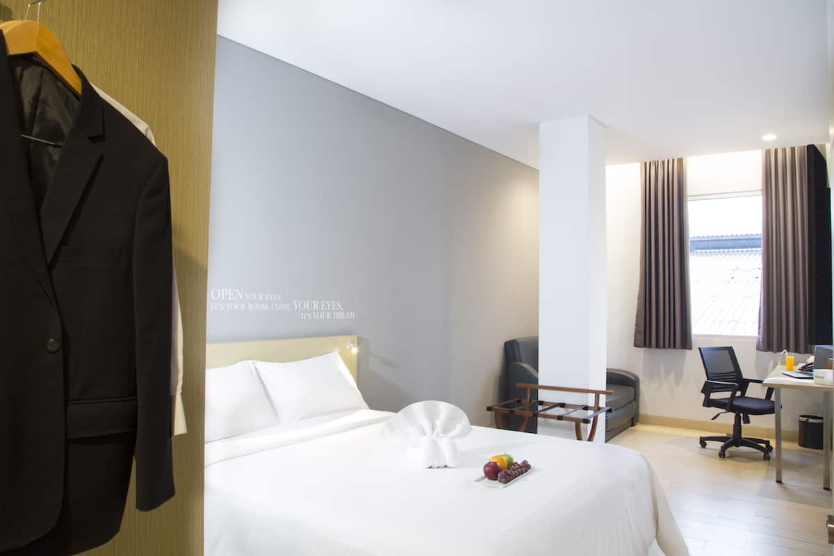 A hotel room with a neatly made bed, a desk with a laptop, a chair, and a coat rack with a jacket.