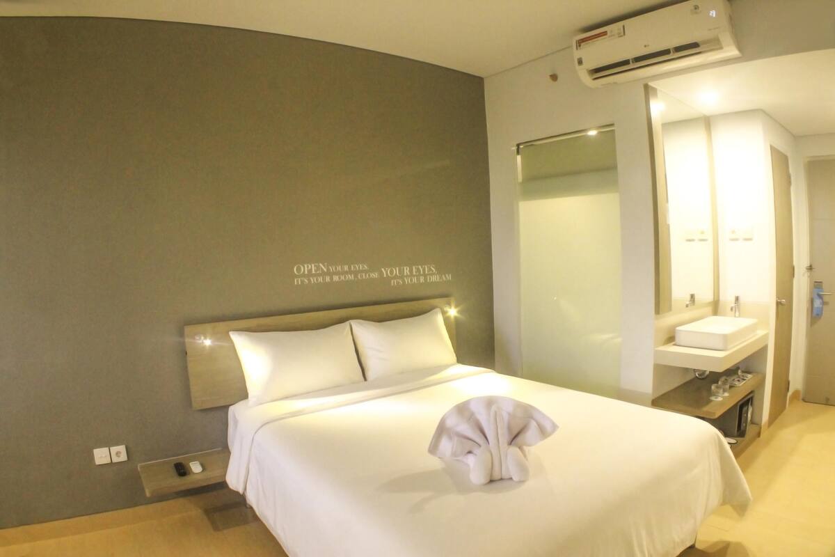 A hotel room with a bed, a bedside table with a remote control, a wall-mounted air conditioner, a bathroom with a sink and mirror, and a motivational quote on the wall.