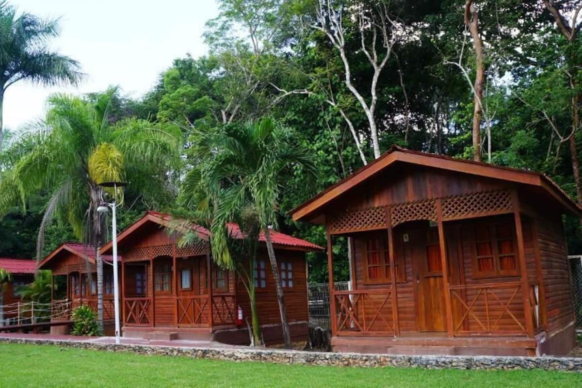 A wooden cabin-style hotel with a porch and multiple windows, surrounded by lush greenery and palm trees.