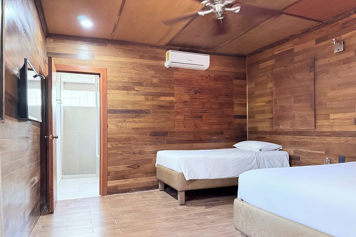 A room with wooden walls, a ceiling fan, an air conditioning unit, two beds, and a door leading to a bathroom.