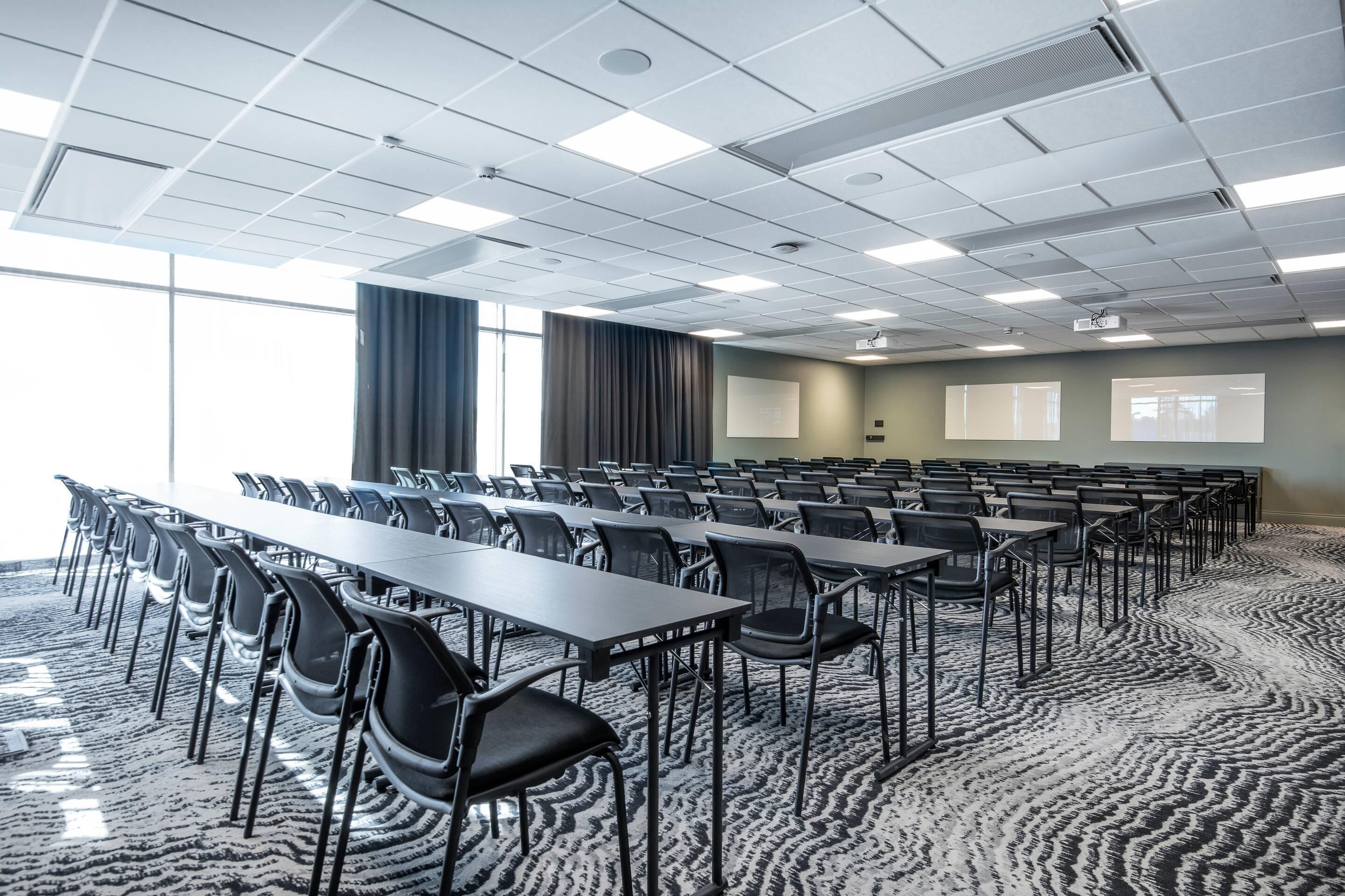 Un salón de conferencias con filas de mesas y sillas, una pantalla de proyector y ventanas amplias.