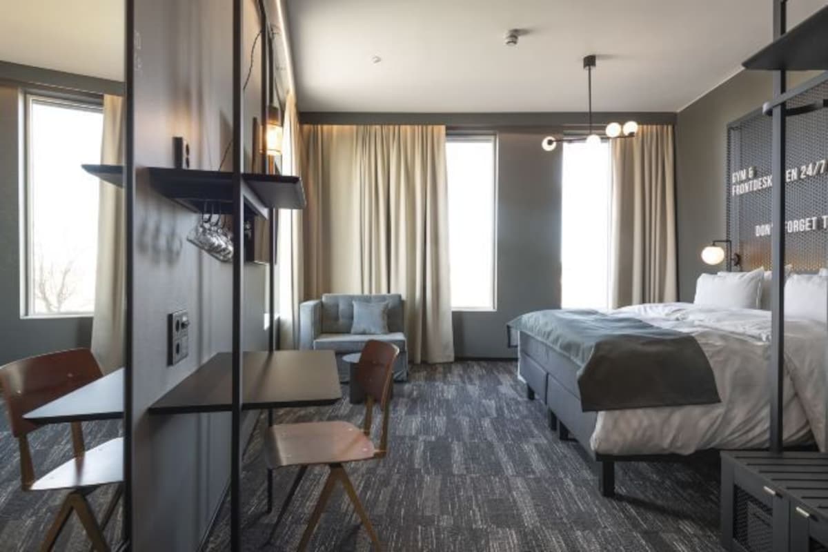 A modern hotel room with a bed, a desk, two chairs, a sofa, and a large window with curtains.