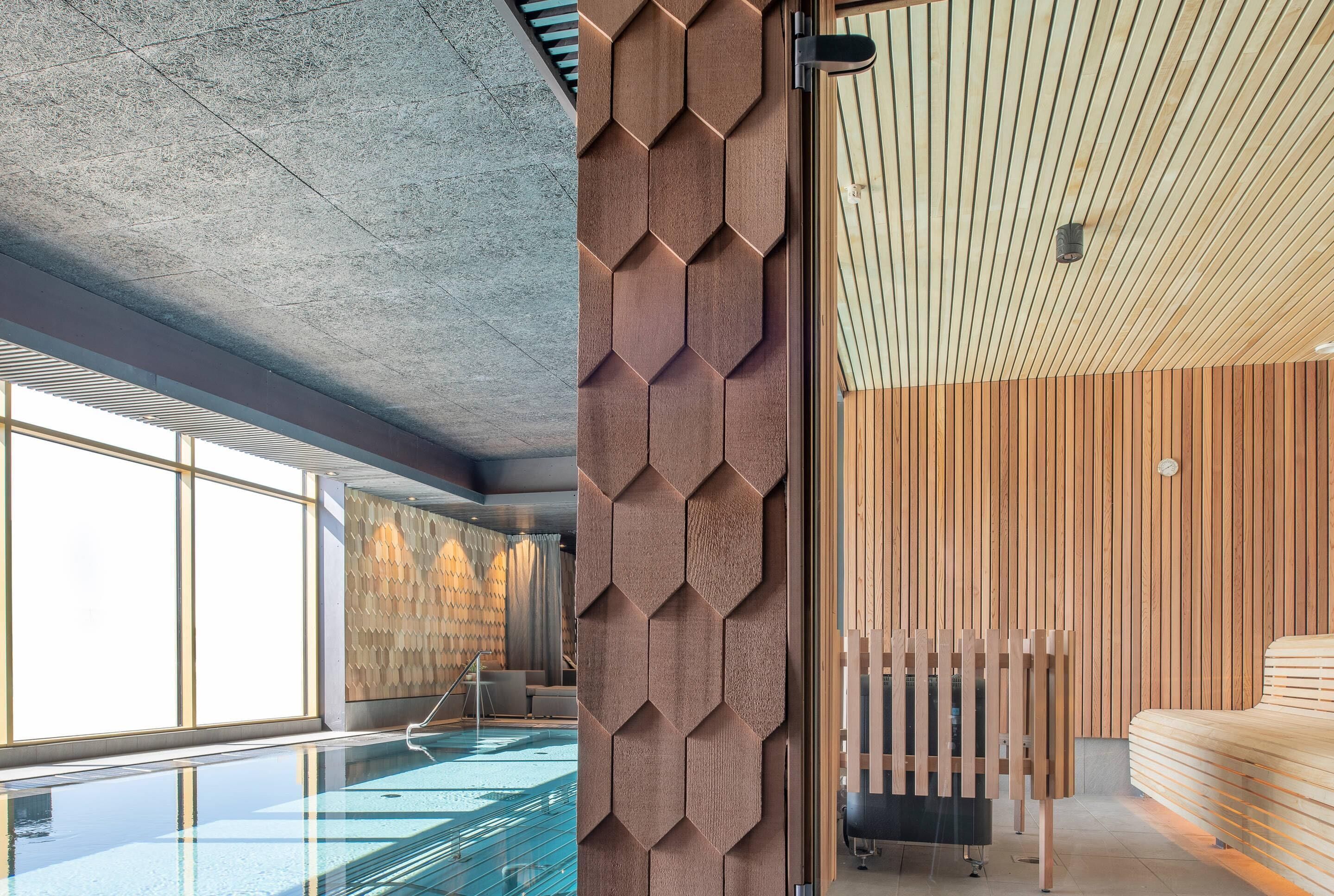 A modern hotel interior with a swimming pool, wooden panel walls, and a ceiling with a geometric pattern.