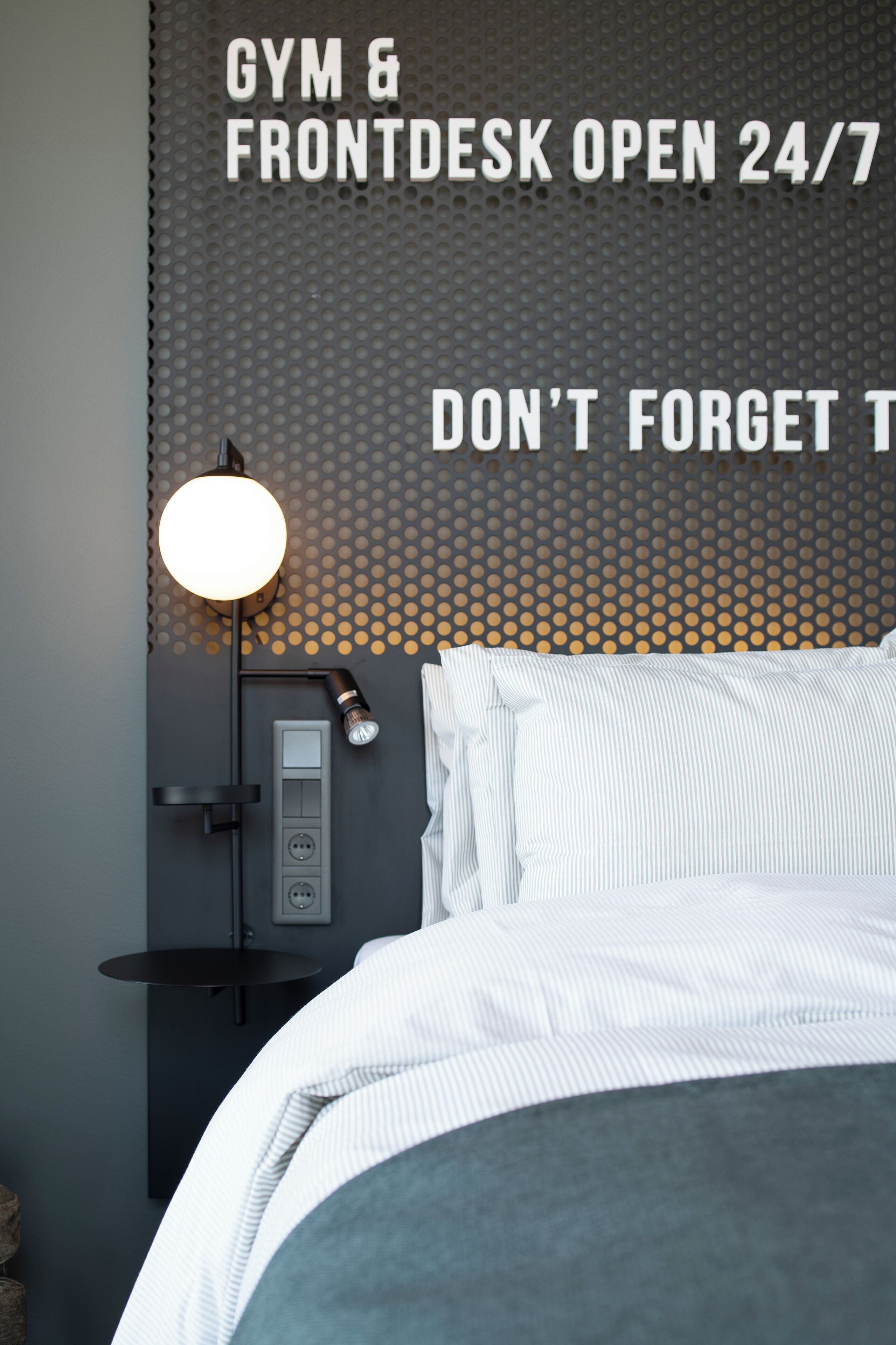 Habitación de hotel moderna con cama, escritorio y lámpara de pared. Un cartel indica que el gimnasio y la recepción están abiertos las 24 horas, todos los días.