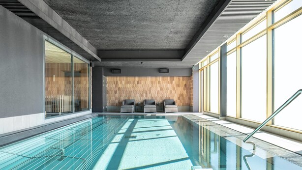 A modern indoor swimming pool with clear glass walls, a wooden wall with geometric patterns, and a seating area with chairs. - Quality Hotel The Box (Linkoping)