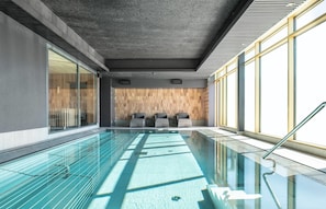 Una piscina interior moderna con paredes de vidrio transparente, un muro de madera con diseños geométricos y un área de descanso con sillas.