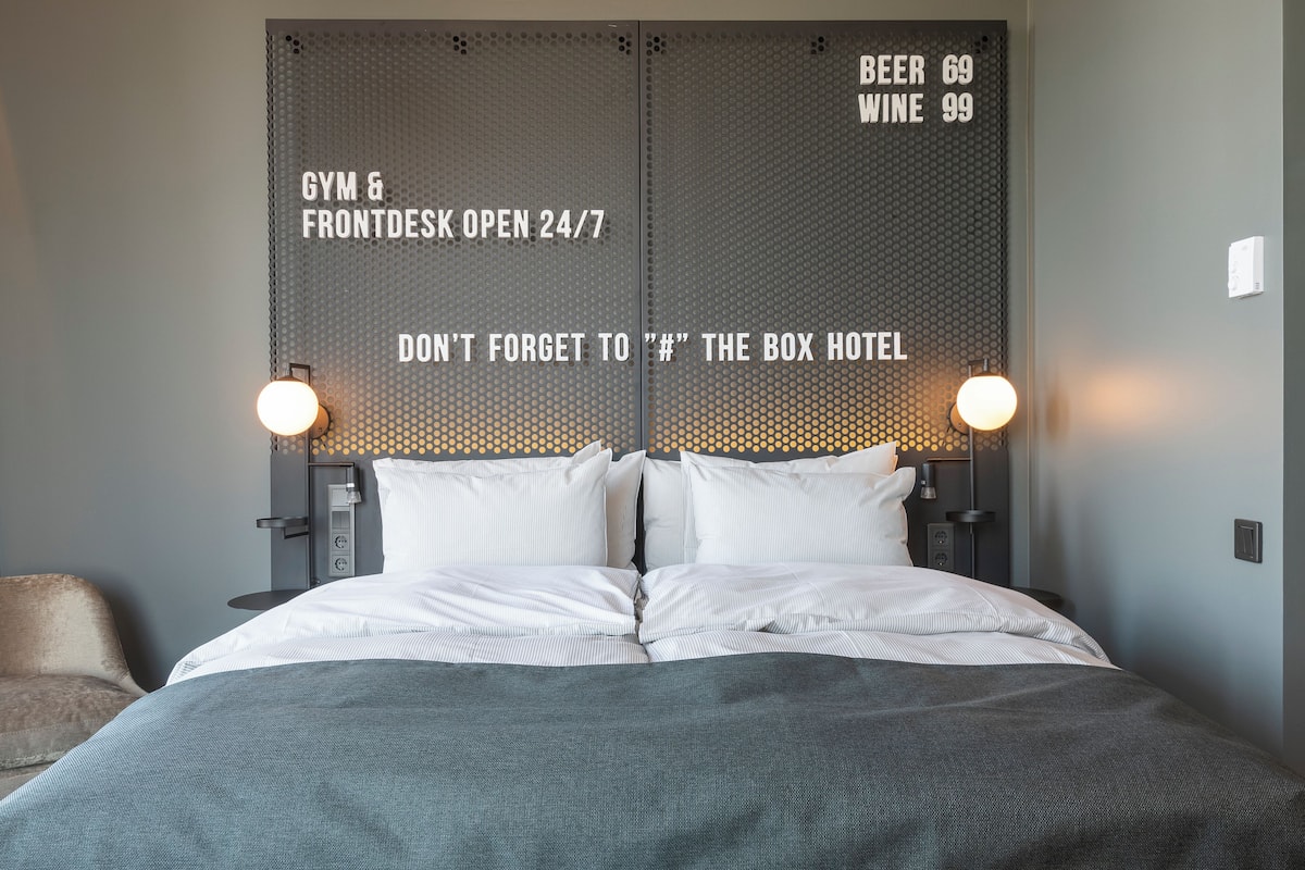 Una habitación de hotel con una cama, dos mesitas de noche y una pared que muestra los servicios, como gimnasio, recepción abierta las 24 horas y caja fuerte del hotel.