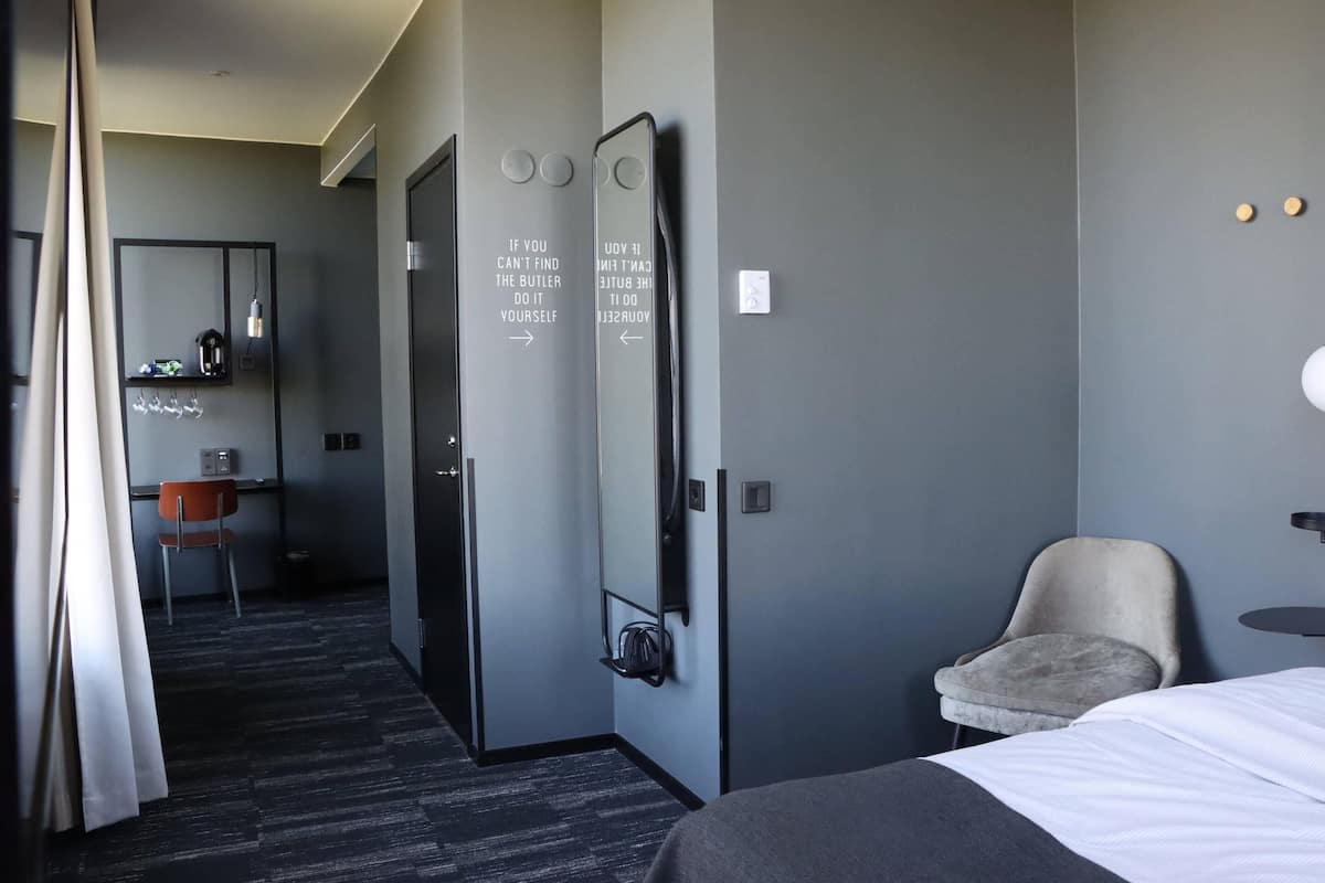 Una habitación de hotel con una cama, una silla, un escritorio con silla, un espejo y una estantería de pared con tetera y tazas.