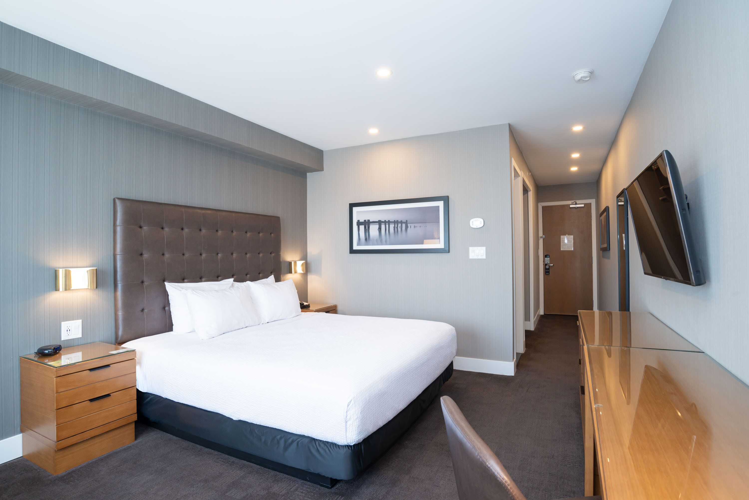 A modern hotel room with a large bed, a bedside table, a flat-screen TV mounted on the wall, and a framed picture above the bed.