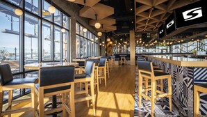 Un restaurante moderno con amplios ventanales, mesas y sillas de madera, y lámparas colgantes.