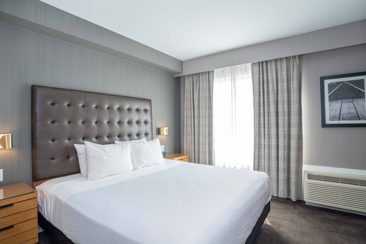 Habitación de hotel con una cama grande, cabecera, mesita de noche, lámpara fijada a la pared, ventana con cortinas e imagen enmarcadas en la pared.