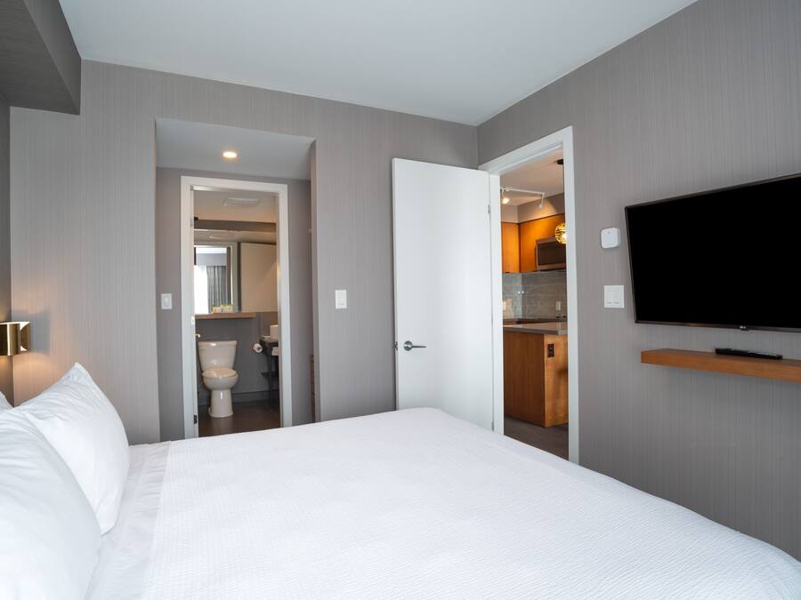 Habitación de hotel con cama, televisor montado en la pared y baño visible a través de una puerta abierta.
