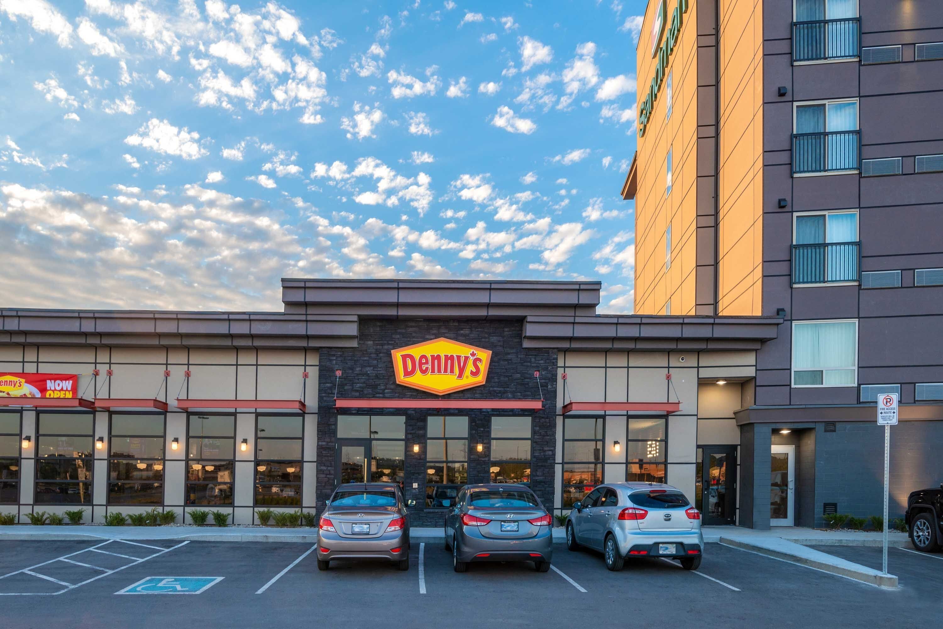 Ein Denny's Restaurant mit drei parkenden Autos vor der Fassade, einem Parkschild und einem bewölkten, sonnigen Himmel.