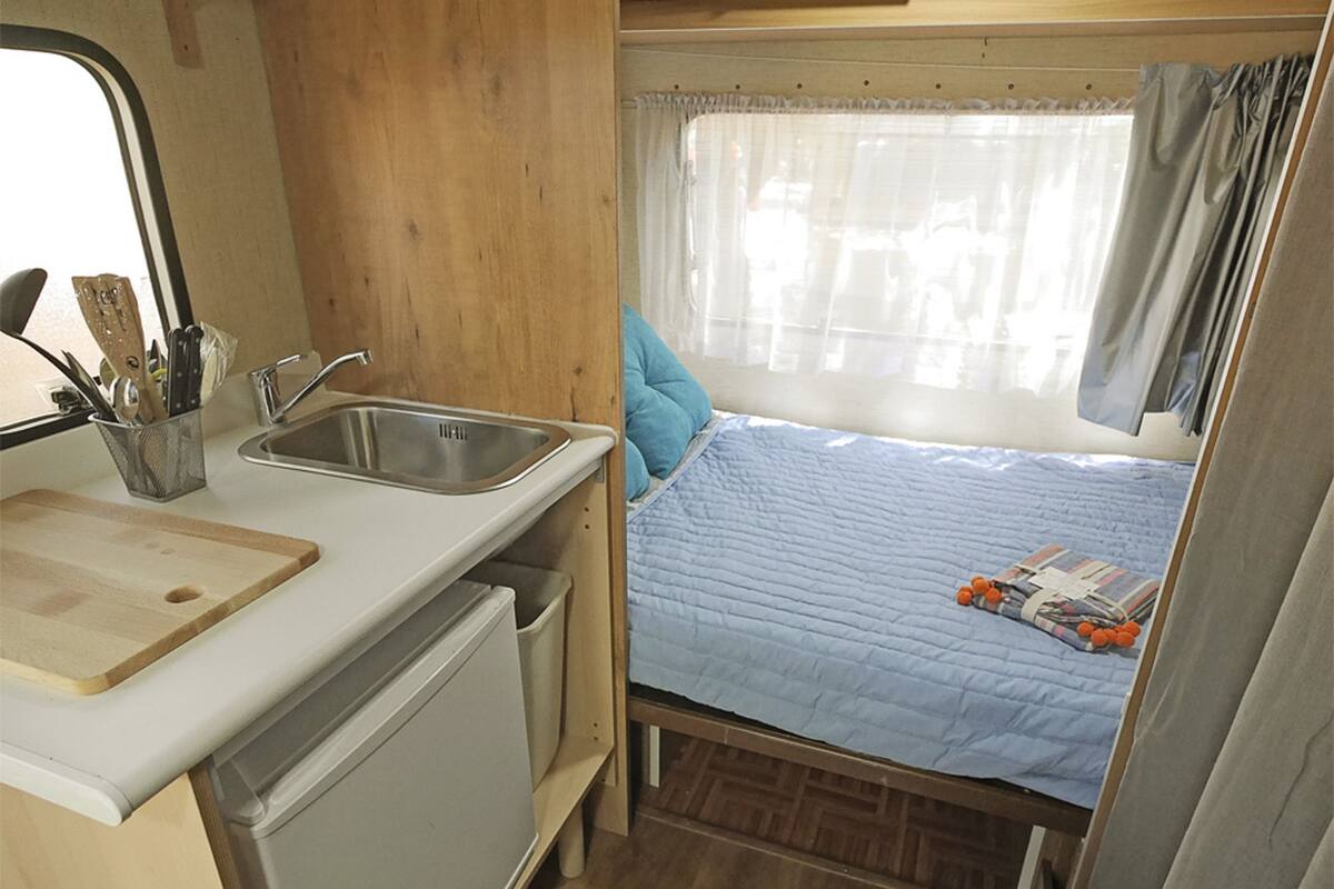 A compact RV interior with a kitchenette, a bed, and a window with curtains.