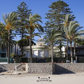 Propiedad frente a la playa con palmeras, un muro de piedra y un edificio de fachada blanca y detalles verdes.