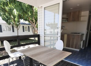 Un área de comedor exterior moderna con una mesa y dos sillas blancas, una terraza de madera y vistas a una casa con un árbol.