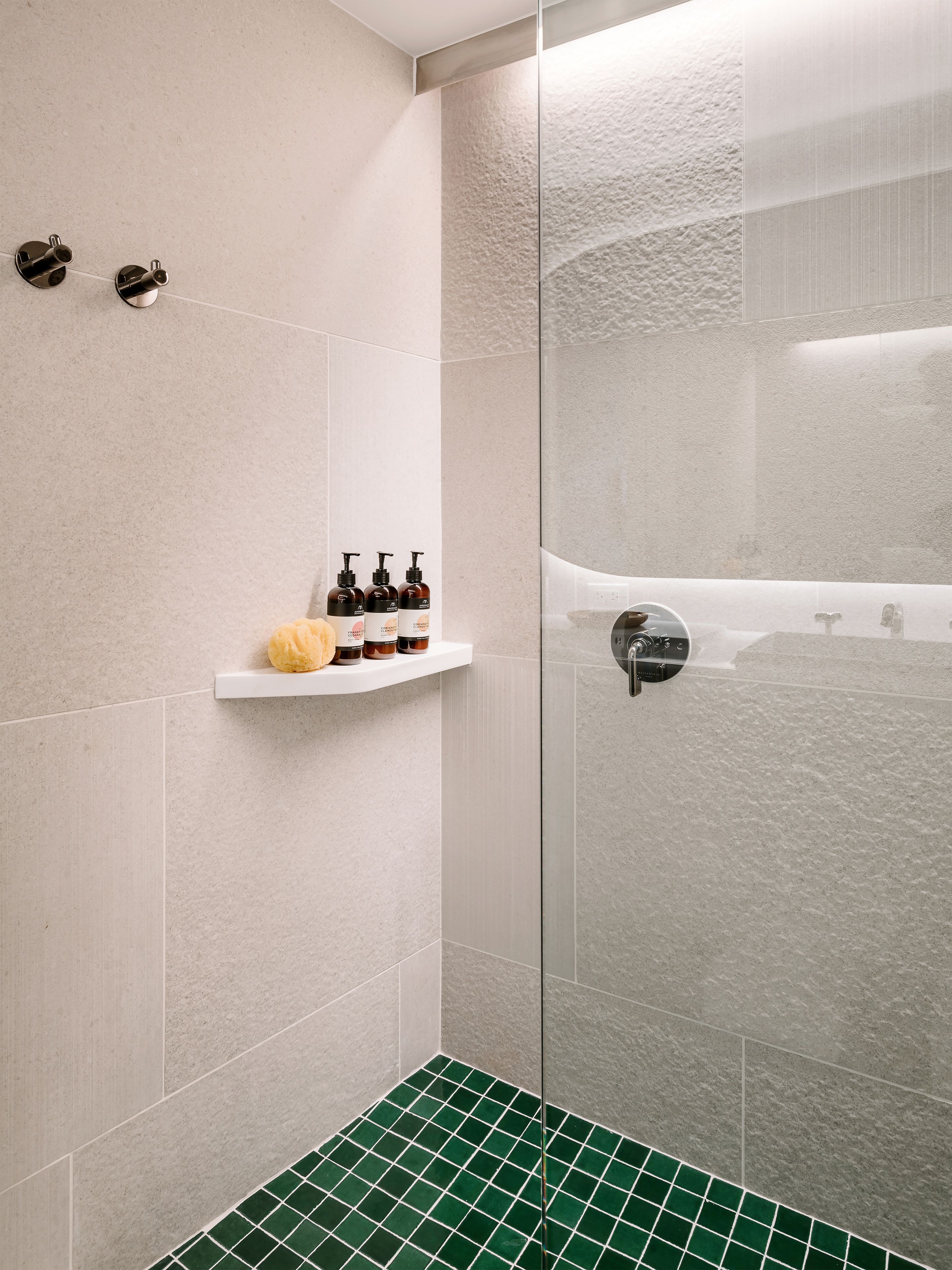 A modern bathroom with a glass shower enclosure, a white shelf with toiletries, and a green-tiled shower floor.
