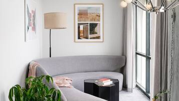 A modern living room with a grey sofa, a side table, a floor lamp, a potted plant, and a large window with curtains.
