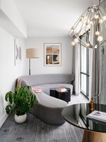 Un salon moderne avec un canapé gris, une table d’appoint, une lampe sur pied, une plante en pot et une grande fenêtre avec des rideaux.