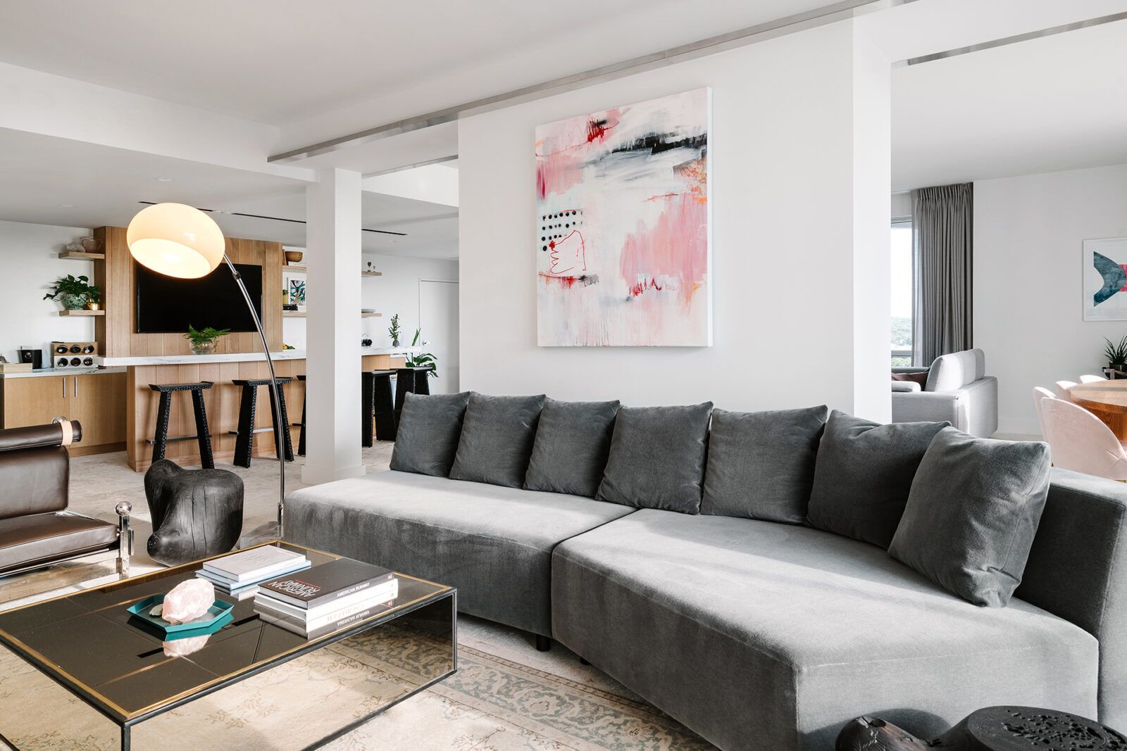 A modern living room with a grey sectional sofa, a coffee table with books, a pendant light, and a large abstract painting on the wall.