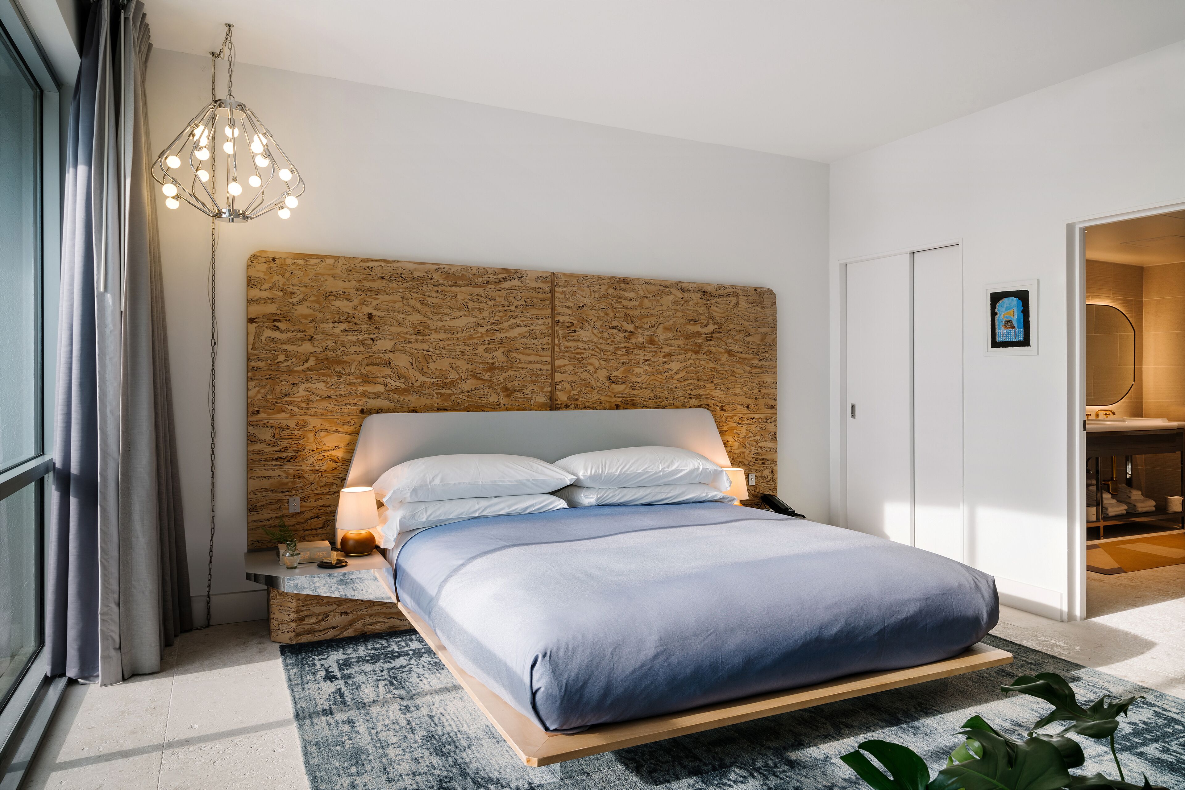 A modern bedroom with a wooden headboard, a bed with a blue mattress, a bedside table with a lamp, and a view of a bathroom through an open door.