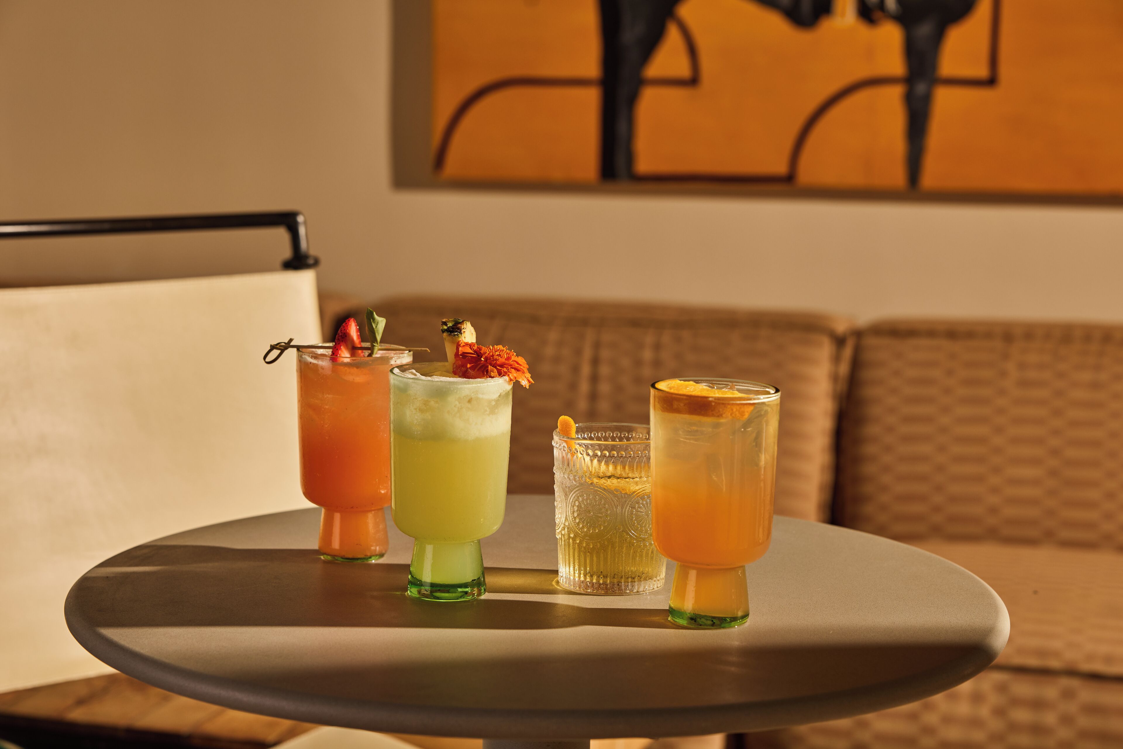 A round table with four drinks, each with a different garnish, in a room with a couch and a painting on the wall.