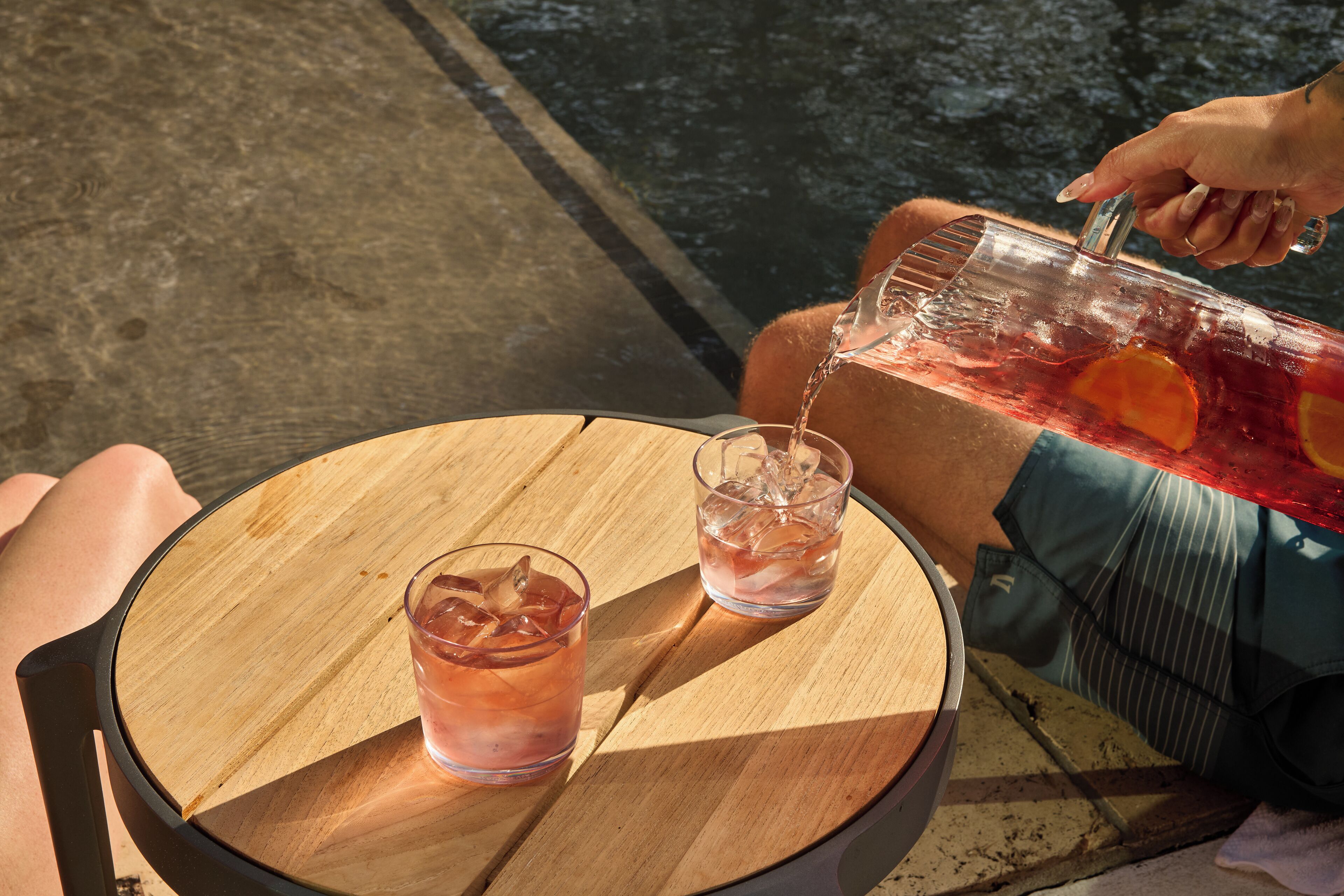 Une personne verse une boisson d’une carafe dans deux verres placés sur une table ronde en bois.