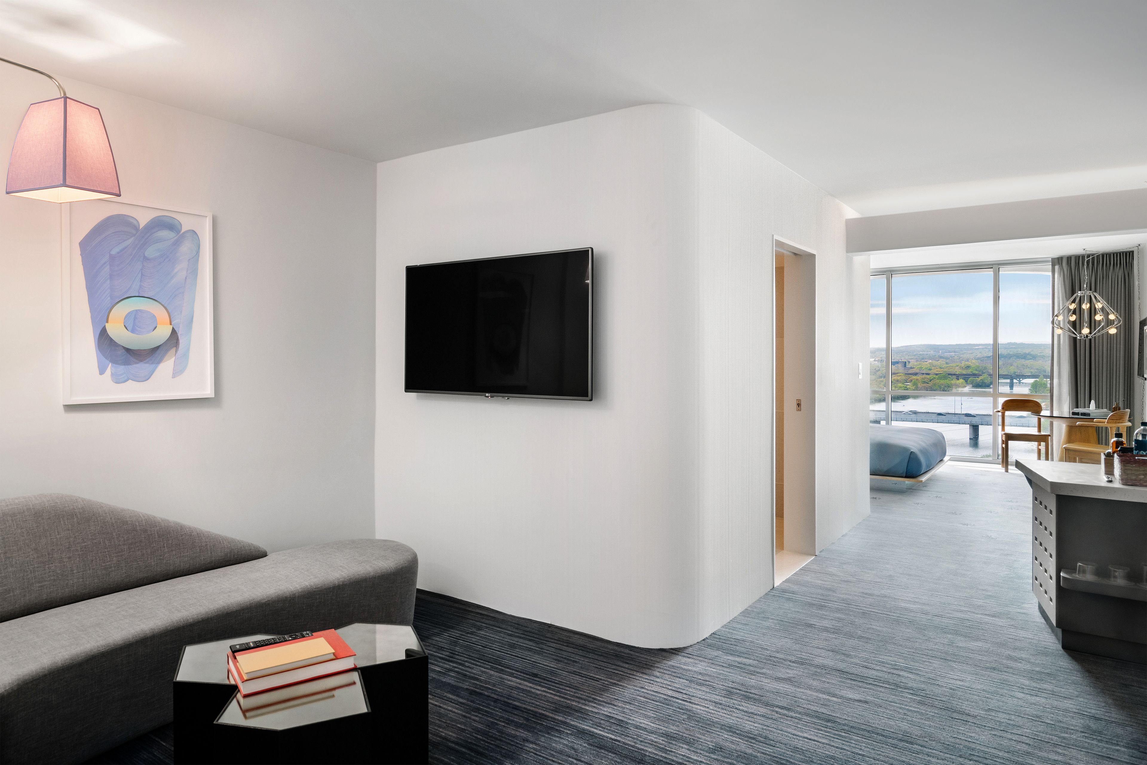 A modern hotel room with a grey sofa, a flat-screen TV, a painting, and a view of the outdoors.