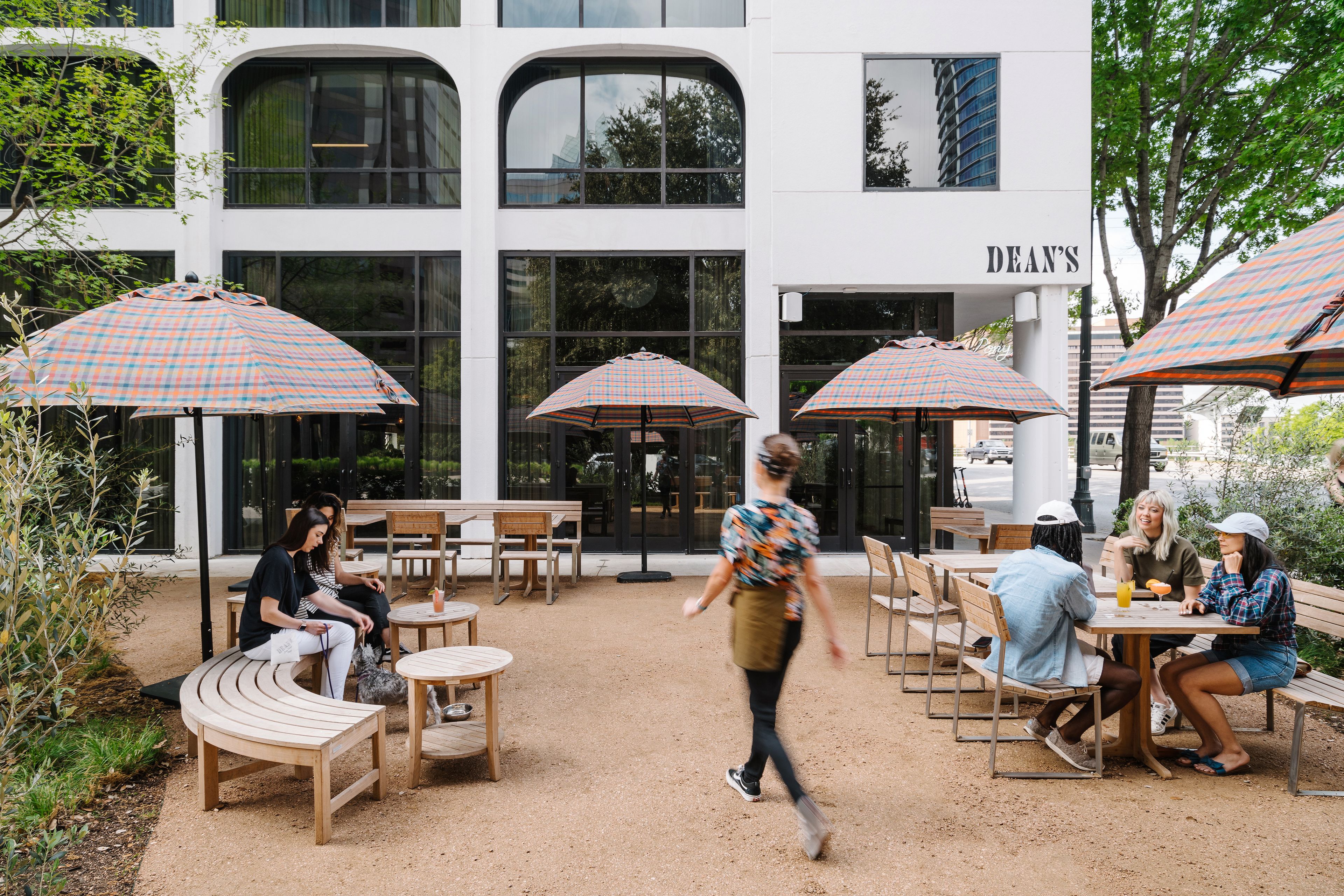 Un área de café al aire libre con mesas, bancos, sombrillas y un edificio con un letrero que dice "DEAN'S".