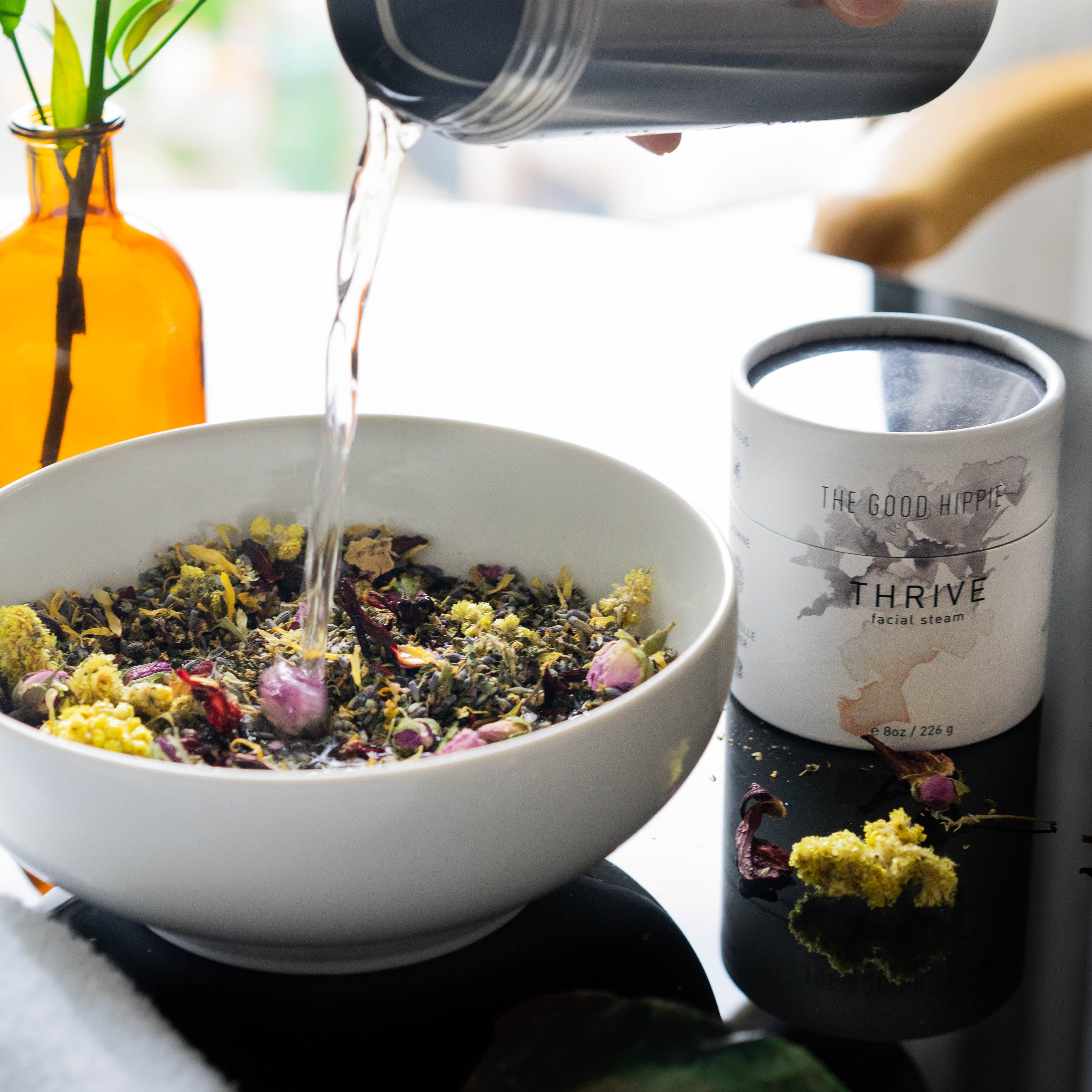 A bowl filled with dried flowers and herbs, a container labeled "THRIVE facial steam," and a clear liquid being poured into the bowl.