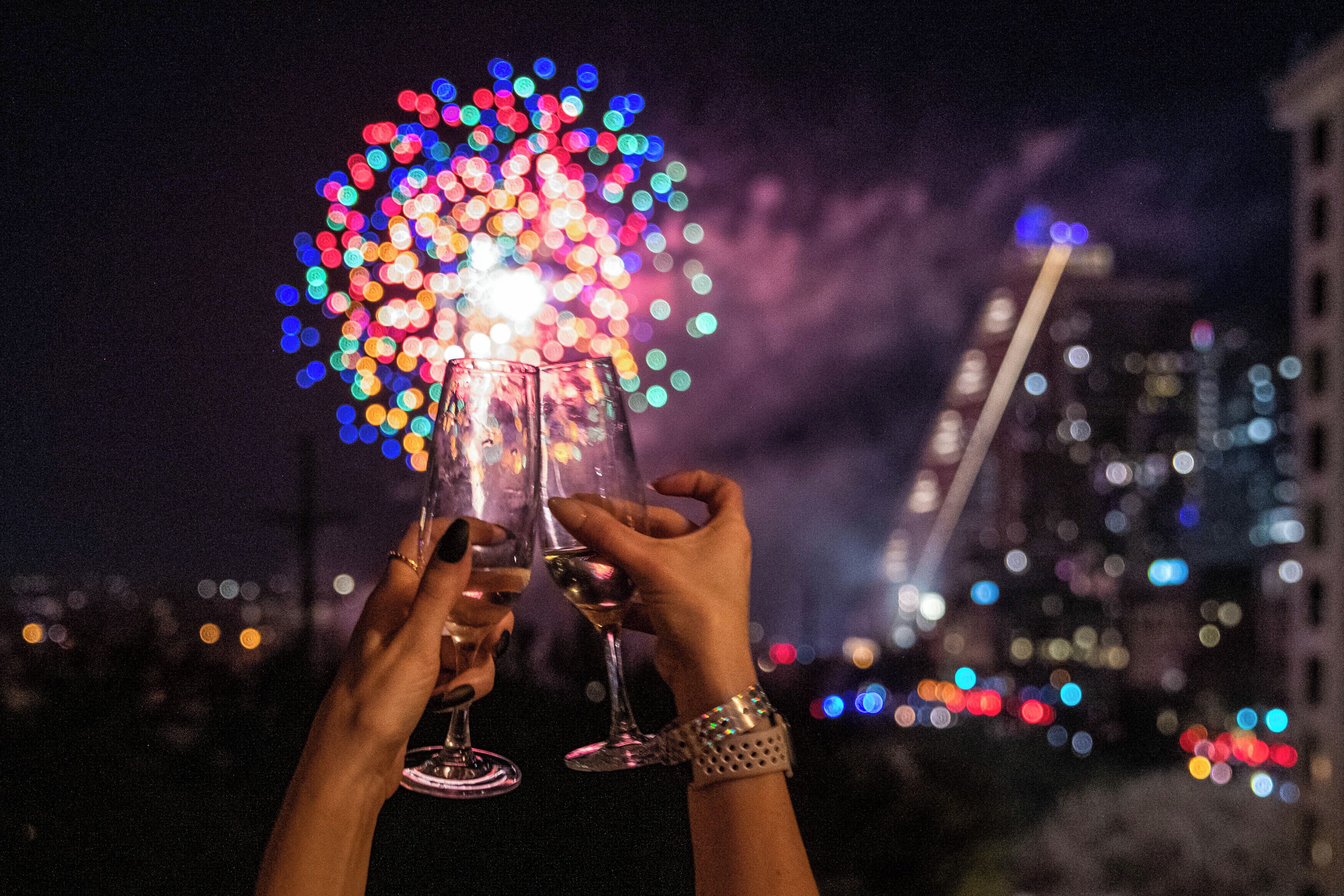 Two hands holding wine glasses with a colorful fireworks display in the background.